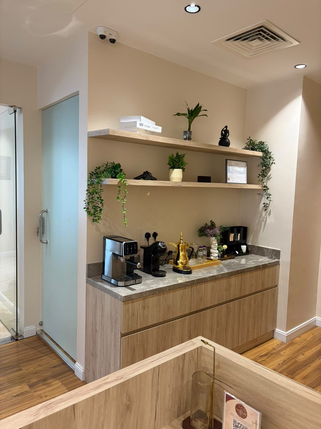 Office pantry counter with light-oak drawers and doors, a grey stone top and floating shelves with plants