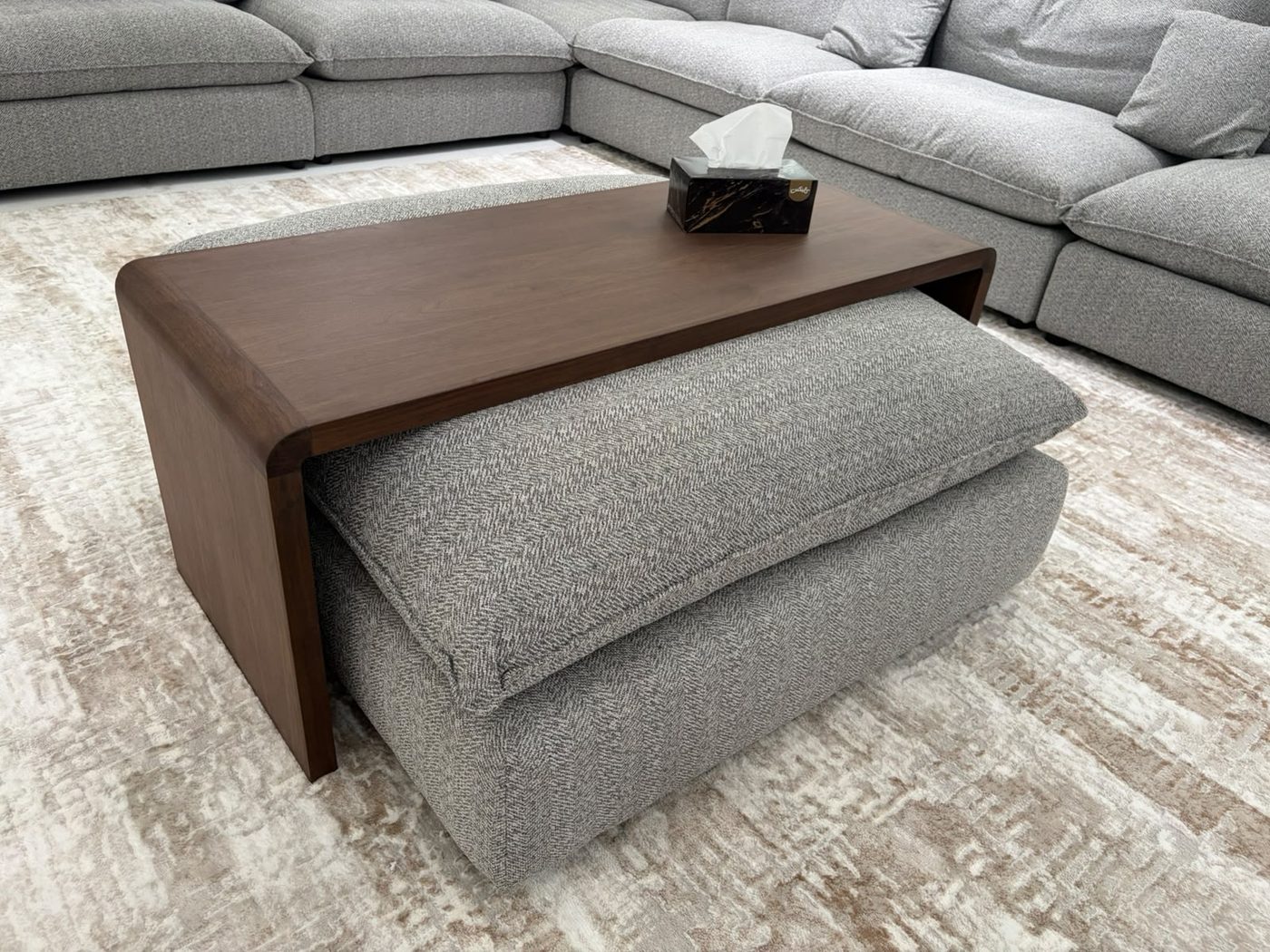 Custom walnut coffee table bridging a grey upholstered ottoman on a textured rug